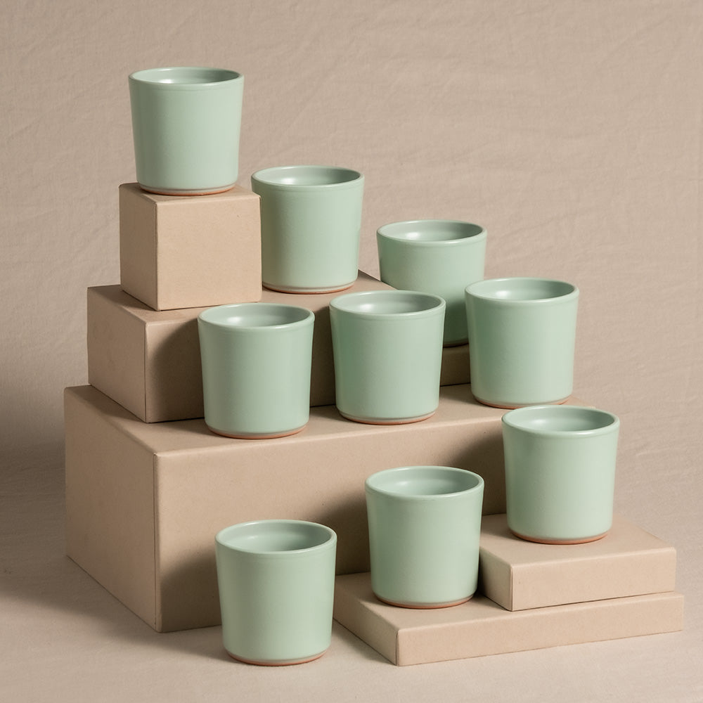 Ein Display mit neun hellgrünen Keramik-Baby-Topfsets 'Sencillo' (9 Stk.) angeordnet auf hellbraunen Kartons unterschiedlicher Höhe vor einem beigen Hintergrund. Die Keramikbecher haben ein schlichtes Design mit einer leicht konischen Form und einem braunen Standrand.