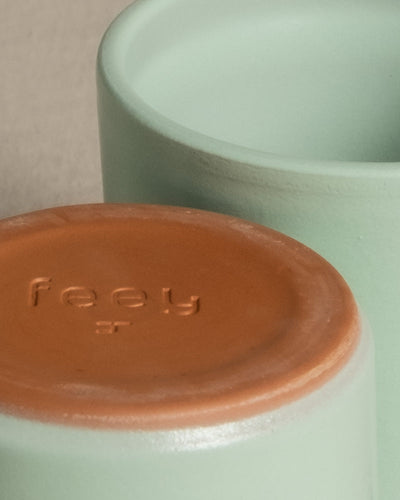 Nahaufnahme von zwei Keramikgefäßen mit schlichtem Design, eines liegt auf der Seite mit dem Boden nach oben, so dass das Wort „feey“ in den braunen, unglasierten Boden geprägt ist. Die übrigen Gefäße sind hellgrün und haben eine glatte Textur. Produktname: Keramik Baby-Topfset „Sencillo“ (9 Stk.)