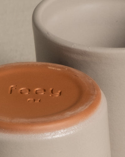 Nahaufnahme des Bodens von zwei zylindrischen Keramik-Baby-Topfsets 'Sencillo' (9 Stk.). Die Töpfe bestechen durch ein schlichtes Design, hellgrau mit glatter Textur. Der Boden eines Topfes hat eine orange-braune Oberfläche, in die das Wort „feey“ und ein kleines Logo eingeprägt sind. Der Hintergrund ist neutral und unscharf.