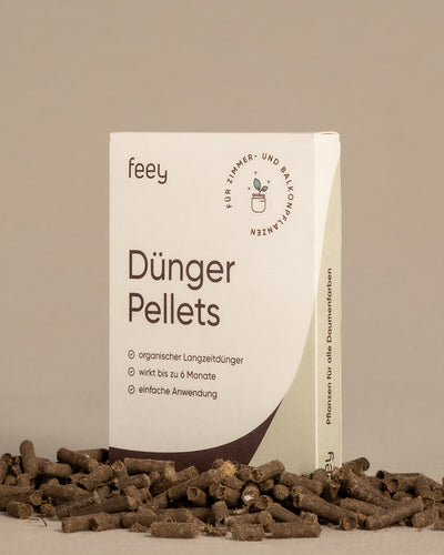 Abgebildet ist eine Schachtel „Biodünger Pellets“ von feey, in der braune Düngerpellets verstreut sind. Die Verpackung wirbt für den organischen Langzeitdünger aus Schafwolle, der für Zimmerpflanzen und Balkonpflanzen geeignet ist, bis zu 6 Monate wirksam ist und einfach zu handhaben ist. 