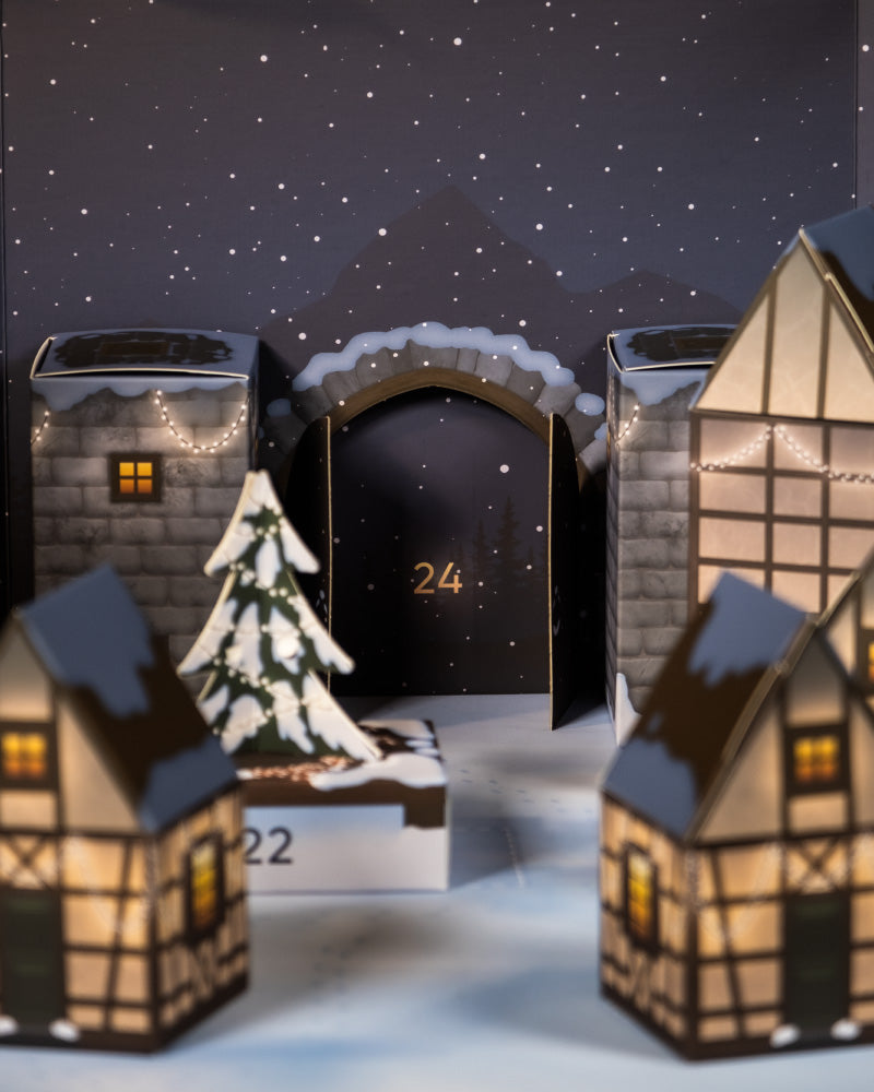 Eine festliche Weihnachtsszene zeigt ein kleines Dorf, das mit Lichtern und echten Pflanzen als Dekoration geschmückt ist. Im Hintergrund dient eine Tür mit der Aufschrift „24“ als Pflanzen-Adventskalender vor einem schneebedeckten Berg, mit einem geschmückten Weihnachtsbaum und schneebedeckten Häusern im Vordergrund.