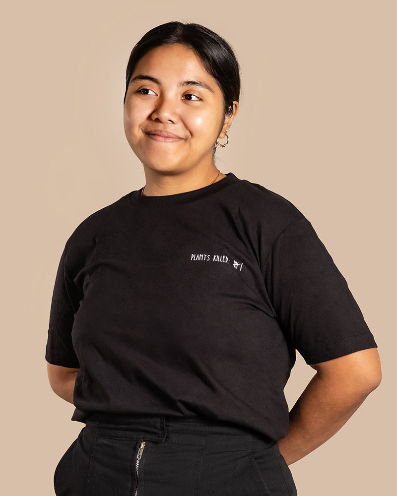 Eine Person steht vor einem beigen Hintergrund, lächelt leicht und blickt aus dem Bild. Sie hat dunkles Haar zurückgebunden und trägt ein schwarzes T-Shirt aus Plant Shirts mit einem kleinen Text auf der Brust, auf dem in weißer Schrift „PLANTS KILLED MY FAMILY“ steht.