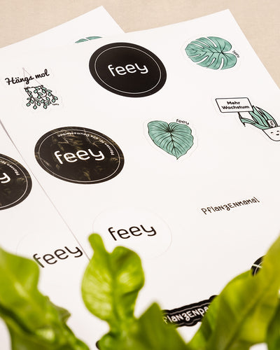 Auf einem Tisch liegen zwei Stickerbögen mit verschiedenen Pflanzenmotiven. Auffällige Sticker sind das Wort „feey“ in verschiedenen Schriftarten, ein Bild einer Topfpflanze mit „Mehr Wachstum“ und eine Sprechblase mit „Hänga mol“. Im Vordergrund sind grüne Blätter zu sehen, perfekt für kreative Köpfe.