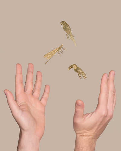 Zwei Hände liegen mit den Handflächen einander zugewandt vor einem beigen Hintergrund. Zwischen den Händen schweben anmutig drei goldene, minimalistische Illustrationen – ein Papagei, eine Heuschrecke und ein Tukan. Diese einzigartige Anordnung unterstreicht den Charme der dekorativen Tierischen Pflanzenhänger, die oft in Deko für Pflanzen-Kollektionen verwendet werden.