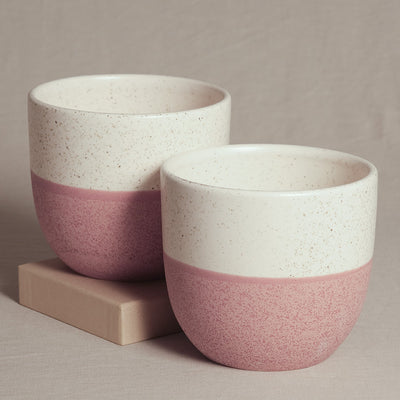 Zwei handgefertigte Keramiktöpfe, das Keramik Topfset 'Variado' (2 × 14), mit minimalistischem Design. Beide Töpfe haben eine weiße obere Hälfte mit braunen Sprenkeln und eine rosa untere Hälfte, wodurch ein eleganter Kontrast entsteht. Perfekt für Zimmerpflanzen, stehen sie auf einem beigen, kartonähnlichen Block mit einem weichen, neutralen Hintergrund.