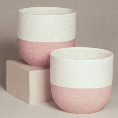 Zwei handgefertigte Keramik-Topfsets „Variado“ (2 × 18) mit gesprenkeltem weiß-rosa Design werden ausgestellt. Eines steht auf einem beigen Sockel, etwas höher als das andere, wodurch ein optisch ansprechendes versetztes Arrangement vor einem neutralen Hintergrund entsteht.
