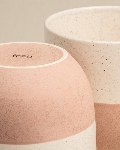 Es werden zwei Keramiktassen mit gesprenkelter Textur gezeigt. Eine Tasse mit einem rosa-weißen Farbverlauf steht verkehrt herum und zeigt das Wort „feely“ auf ihrem Boden. Das andere handgefertigte Keramik-Topfset „Variado“ (3 × 16) mit einem weiß-beigen gesprenkelten Design steht aufrecht hinter dem ersten Keramiktöpfe.