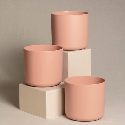 Drei rosafarbene, zylindrische Keramiktopfsets (3 x 14) in unterschiedlichen Höhen werden auf und um zwei gestapelte rechteckige weiße Blöcke vor einem neutralen Hintergrund ausgestellt. Das Arrangement ergibt eine optisch ansprechende und minimalistische Komposition, ideal für die Präsentation von Zimmerpflanzen.