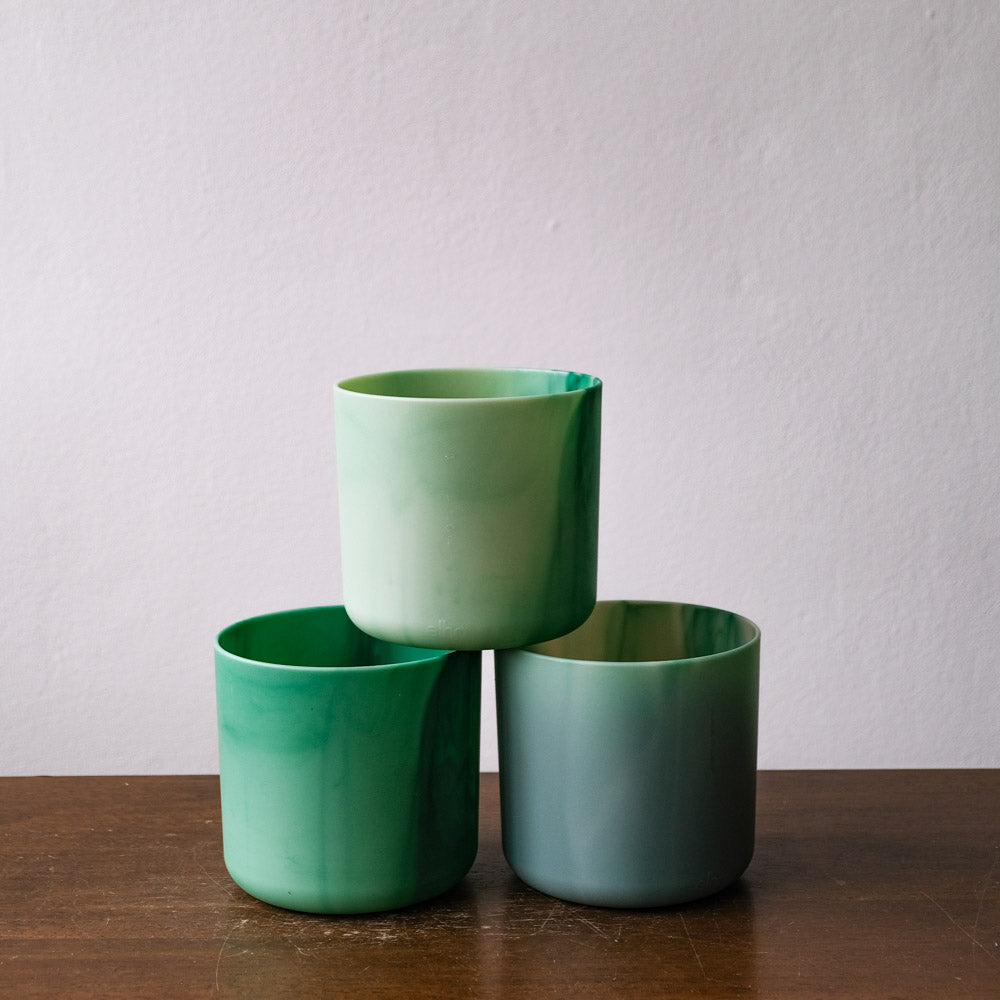 Drei Keramiktassen, dreieckig gestapelt. Die obere Tasse ist hellgrün, während die unteren Tassen dunkelgrün bzw. blaugrün sind. Sie ähneln einem Miniatur-Topfset (3 x 14) und stehen auf einer Holzoberfläche vor einem schlichten, hellen Hintergrund.