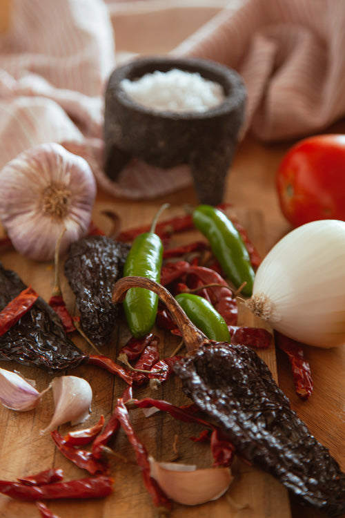 Verschiedenfarbige Chilis auf einem Holzbrett, manche gedörrt, manche nicht, dazwischen ganze und teils geschälte Zwiebeln