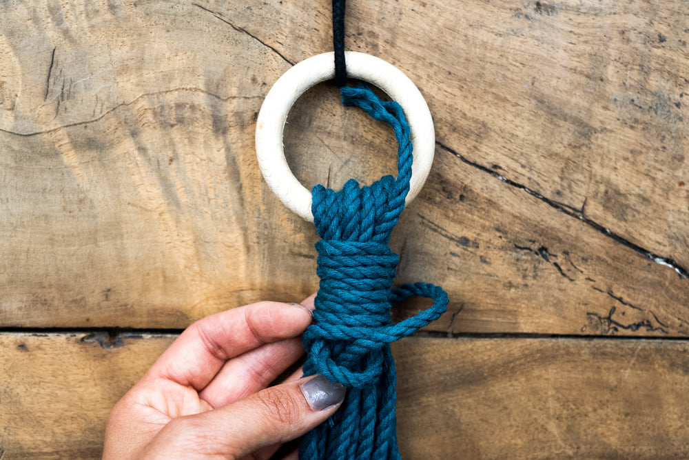 Bild für die Anleitung eines Wickelknoten mit Ring. Die Schnüre sind türkis und liegen auf einem Holztisch.