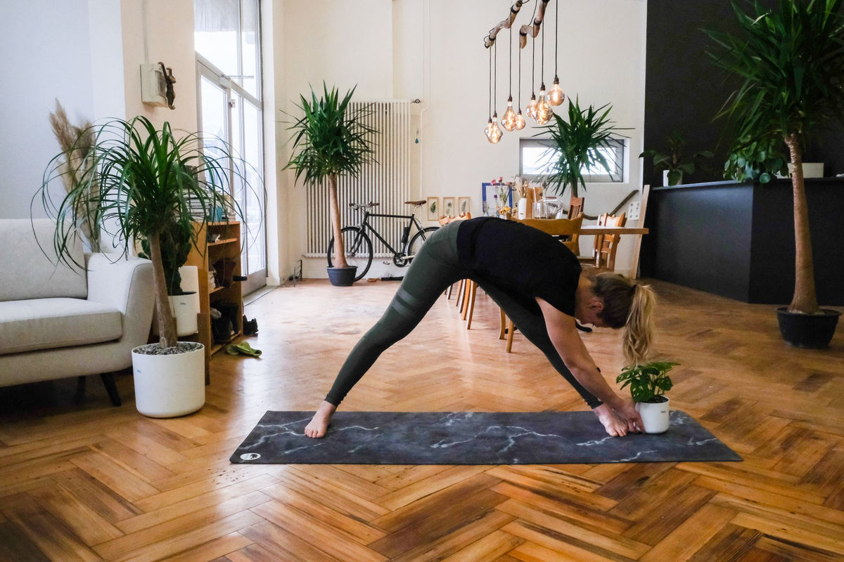 Plant-Yogi Hanna steht mit gestreckten, breiten Beinen über ihr linkes Bein gebeugt, die Hände mit der Pflanze am Boden