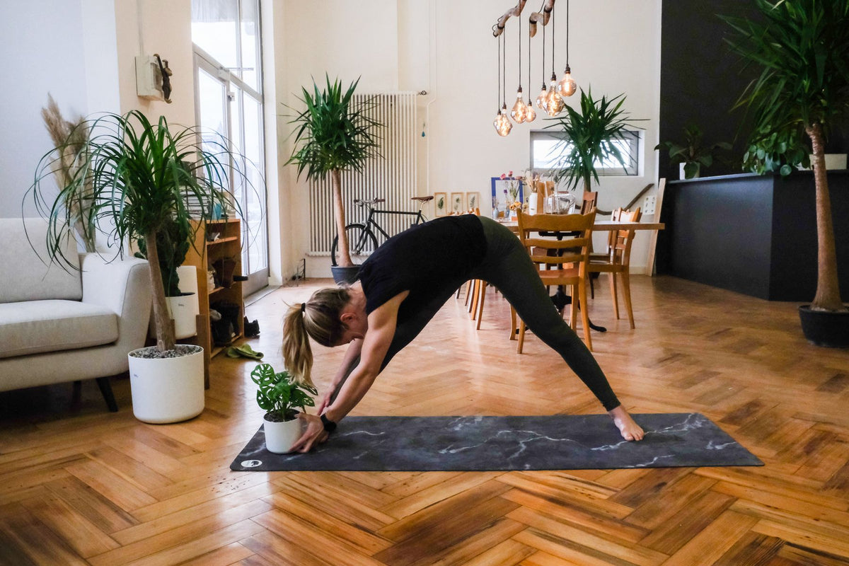 Plant-Yogi Hanna steht mit gestreckten, breiten Beinen über ihr rechtes Bein gebeugt, die Hände mit der Pflanze am Boden