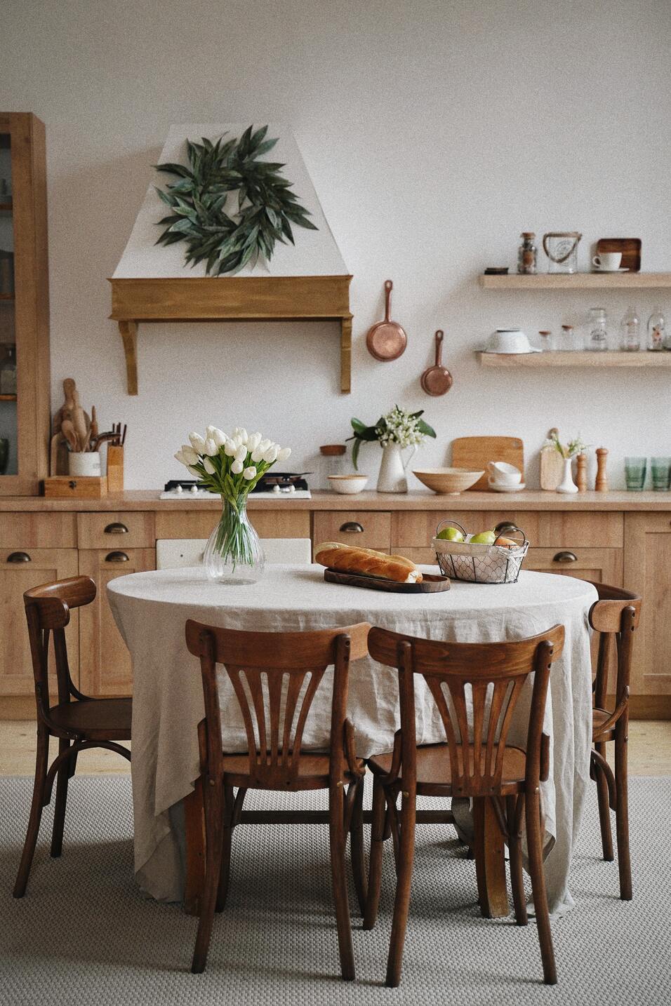 Holzstühle um einen weiss gedeckten, runden Tisch, darauf ein Blumenstrauss und hinten auf der hölzernen Anrichte eine Pflanze in der Vase