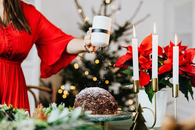 Frau in rotem Kleid streut Puderzucker über einen Kuchen auf einer festlich gedeckten Tafel, hinter dem Kerzenständer mit drei Kerzen ein Weihnachtsstern mit roten Blättern