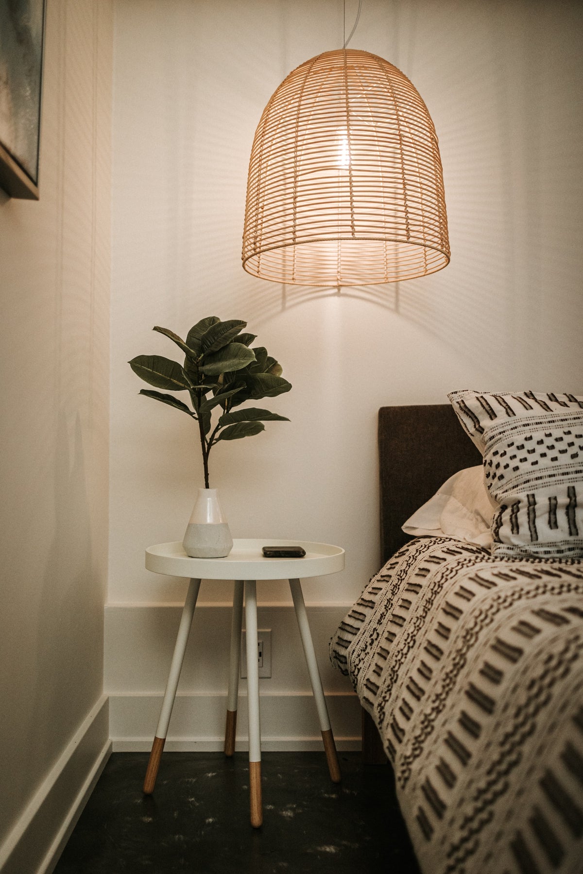 Von der Decke hängt eine grosse Lampe aus Bast. Auf einem weissen Nachttisch steht eine Vase mit einem Ast eines Gummibaums drin. Das Bett neben dem Nachttisch hat schwarz-weiss gemusterte Bettwäsche.